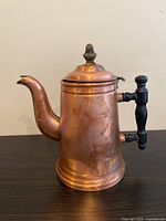 Side angle of a tall copper kettle showing the wooden handle and curved spout, placed on a dark wooden surface against a plain wall.