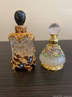 Two glass bottles displayed on a flat surface, showing the ornate metal overlays and stoppers, with clear Versailles bottle on left and frosted bottle on right.