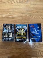 Three hardcover thriller novels on hardwood floor: two Jack Reacher books by Lee Child and one thriller by Karin Smirnoff