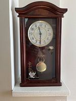 Front view of Masterpiece Quartz wall clock showing wood frame, clock face, pendulum, and floral glass decoration.