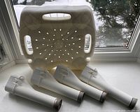 Disassembled white plastic heavy duty shower seat with four legs placed on windowsill, viewed from above showing drainage holes and hand grips