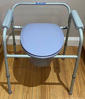 Front view of Probasics three-in-one commode with grey padded arms and plastic seat with lid on mahogany floor background.