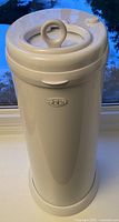 White cylindrical Ubbi steel diaper pail with rounded top and circular sliding lid with handle, standing on a window sill.