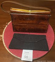 Photo of both vintage purses shown on a round red placemat: a brown snakeskin faux handbag with gold clasp and a black evening clutch with embossed floral design.