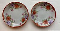 Top down image of two small round ceramic saucers with floral artwork in orange, purple and pink, each with gold rim detail