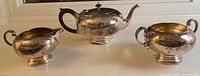 Photo showing entire Victorian plated tea set with teapot, creamer, and sugar bowl arranged in a row on a white surface.