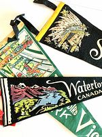 Close-up photo of three vintage souvenir pennants highlighting detailed graphics: Thunderbird Park with Haida totems, Waterton Lakes with mountain and lake scene, and St. Thomas with First Nations man in headdress.