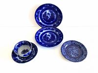 Five early flow blue dishes shown arranged on a white background: two stacked matching plates, one smaller floral plate, a deep tea saucer with cup on it, and a handleless cup.