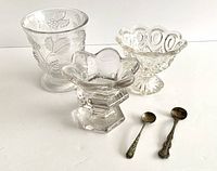 Pressed glass spooner, two master salt cellars, and two salt spoons arranged on a white surface.