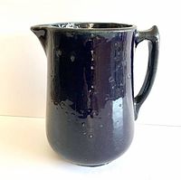Front view of a tall, heavy stoneware pitcher with a dark pock-marked blue glaze and a spout on the rim