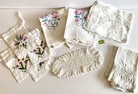 Lot overview showing all 9 items including 4 embroidered floral drawstring bags, crocheted 'Bread' doily, large white placemat with crossed utensils embroidery, guest towel with floral embroidery and openwork, and 2 Angie Strauss tea towels.
