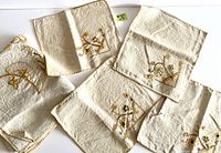Raw linen embroidered napkins or placemats arranged side by side, showing detailed floral embroidery in marigold and black thread.