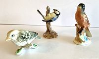 Three vintage ceramic bird figurines shown together on white surface: Goebel bird hanging upside down, finch bird standing, squat bird on tripod base.