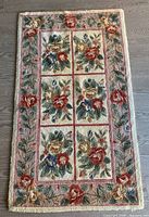 Full view of rectangular handmade wool rug with floral pattern and pink border surrounding six floral panels on cream background.