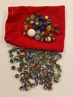Overview photo showing a red drawstring pouch holding some marbles and many glass marbles spread out beneath it on a flat surface. Marbles display multiple colors and swirling patterns.