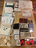Overview of vintage and new embroidered teatowels, napkins, rectangular tablecloths, several sets of vintage coasters, pottery napkin rings, and small boxed linens on a speckled countertop.