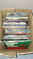 Photo of cardboard box filled with numerous 45RPM records in picture sleeves, viewed from above showing record spines and sleeves.