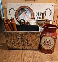 Photo showing blacksmith sign board with horseshoes and text, along with various decorative items in front including framed plate, brass and wood magazine rack, painted milk can, and brass plant pot.