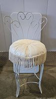 Front view of vintage white metal framed vanity chair with tufted cream cushion and fringe trim showing some staining.
