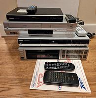 Stacked vintage electronic devices including Zenith VCR, Toshiba DVD/VCR combo player, Toshiba DVD player, and Toshiba VCR with remote controls and documents.