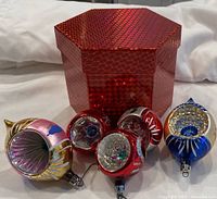 Five vintage glass Christmas ornaments of various colors and styles arranged in front of a red hexagonal reflective box on a white background.