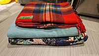 Stack of three folded blankets and quilts showing the red Uruguay wool blanket on top with plaid pattern and fringe edges, the blue wool blanket in the middle, and a floral reversible quilt on the bottom.