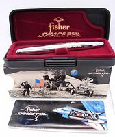 Chrome bullet-shaped Fisher Space Pen inside black hard case with red velvet lining, branded with 'Fisher Space Pen' in gold, placed on original outer box decorated with lunar landing scene and astronaut.