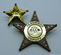 Two vintage metal star-shaped law enforcement badges on a flat surface: larger Deadwood Deputy badge with stagecoach design and smaller Roy Rogers Sheriff badge.