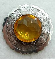 Front view of the round silver kilt pin showing detailed engraved Scottish thistle design around the amber faceted glass stone center.