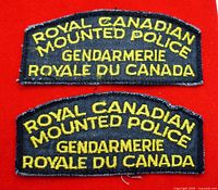 Front view of two Royal Canadian Mounted Police shoulder flashes with yellow embroidery on black fabric, bilingual text, set on red background.