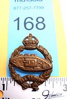 Front view of WWI era Canadian Armoured Corps cap badge showing tank motif, King's Crown, and 'Dear Watch' banner with ruler for scale.