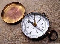 Open view showing interior white dial with compass directions and degrees, black needle, and hinged brass cover open wide on fabric background.