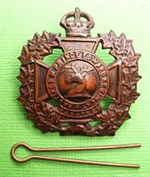 Front view of a metal WWII Hastings and Prince Edward Regiment cap badge on a green background showing detailed maple leaf wreath design and King's Crown.