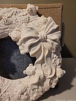 Close-up of the ceramic wreath showing the sculpted teddy bears, pine cones, holly leaves, and a large bow near the bottom, highlighting the unpainted white ceramic surface and detailed texture.