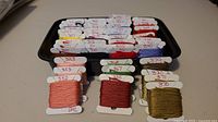 DMC 300 series cross stitch threads neatly arranged on white bobbins inside a black tray, showing color numbers handwritten on each bobbin, colors include shades of red, purple, blue, green, gold, peach.