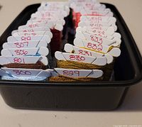 Photo showing DMC 800 series embroidery floss on thread bobbins arranged in rows inside a black plastic container, with numbers written on each bobbin.