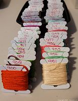 Two images showing multiple DMC 900 series embroidery floss skeins wound on white bobbins with handwritten thread numbers. Colors visible include various shades of green, orange, cream, red, pink, and blue.