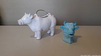 Photo of white pottery cow with a handle and light blue glass bull figurine shown side by side on a surface.