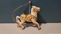 Front view of Lenox porcelain carousel Palomino horse ornament suspended by hanging loop against blue background.