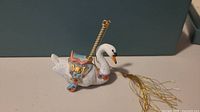 Front-side view of white porcelain swan carousel ornament with detailed saddle and gold hanging string with tassel.