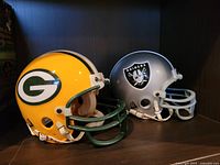 Side view of the two Riddell mini football helmets, Green Bay Packers on the left in yellow and Raiders on the right in silver.