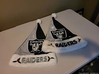 Two Raiders Santa hats with gray/black split design, Raiders patch on one side, white fuzzy brim with 'RAIDERS' embroidery and holly, white pom-pom at top