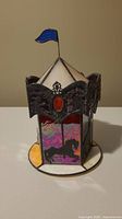 Photo of stained glass carousel light showing partial side view, blue flag on top, horse silhouette in stained glass panels, ornate metal decorations with red accents, circular white base.