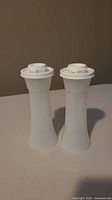Two white Tupperware salt and pepper shakers with screw-on lids placed upright on a table.