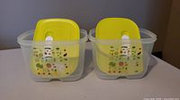 Two small clear plastic Tupperware FridgeSmart containers with yellow lids on a flat surface, showing colorful fruit and vegetable icons on sides.
