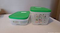 Two translucent plastic Tupperware Fridgesmart containers with green lids, one medium deep and one medium size, side by side on a white surface against a beige wall.