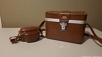 Two brown leather camera cases; one is hard-sided with white stripe and metal clasp, the other is a smaller leather case with strap.
