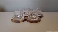 Four textured clear whiskey glasses placed on four round wooden coasters on a plain tabletop.