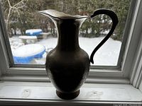 Brass jug with a polished brass body and black curved handle, photographed on a window sill with a snowy outdoor background.