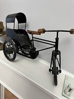 Black metal tricycle shaped decor piece with wooden handles and small cargo bench seat displayed against a white wall.